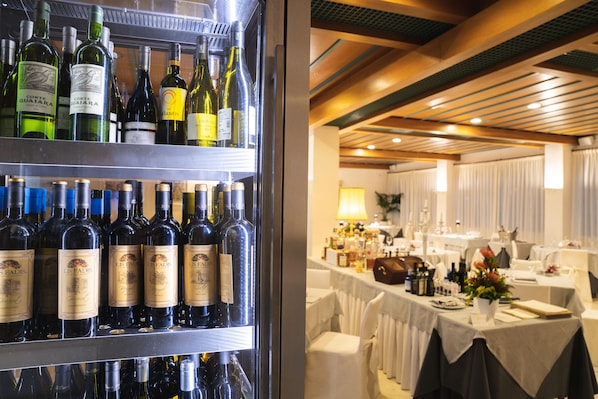 Un refrigerador de vinos repleto de diversas botellas, un comedor con mesas dispuestas para invitados, y un techo con vigas de madera.