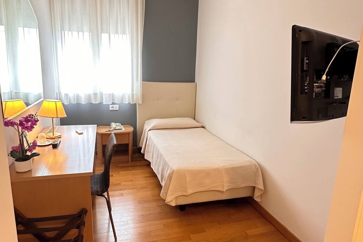 A small, well-lit room with a wooden floor, a desk with a lamp, a bed, a chair, and a TV mounted on the wall.