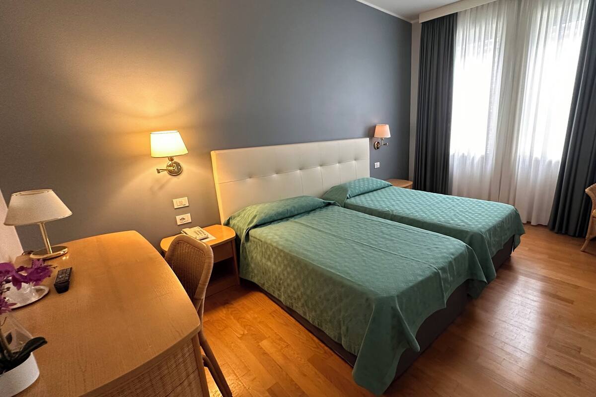 A hotel room with two beds, a desk with a lamp, a chair, and a window with curtains.