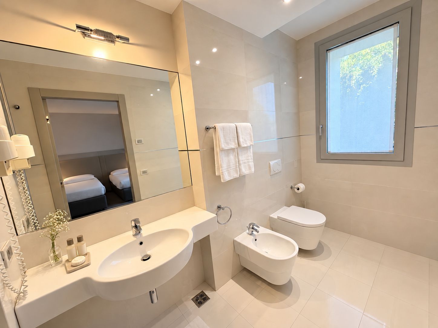A modern bathroom with a large mirror, a double sink, a bidet, and a toilet.