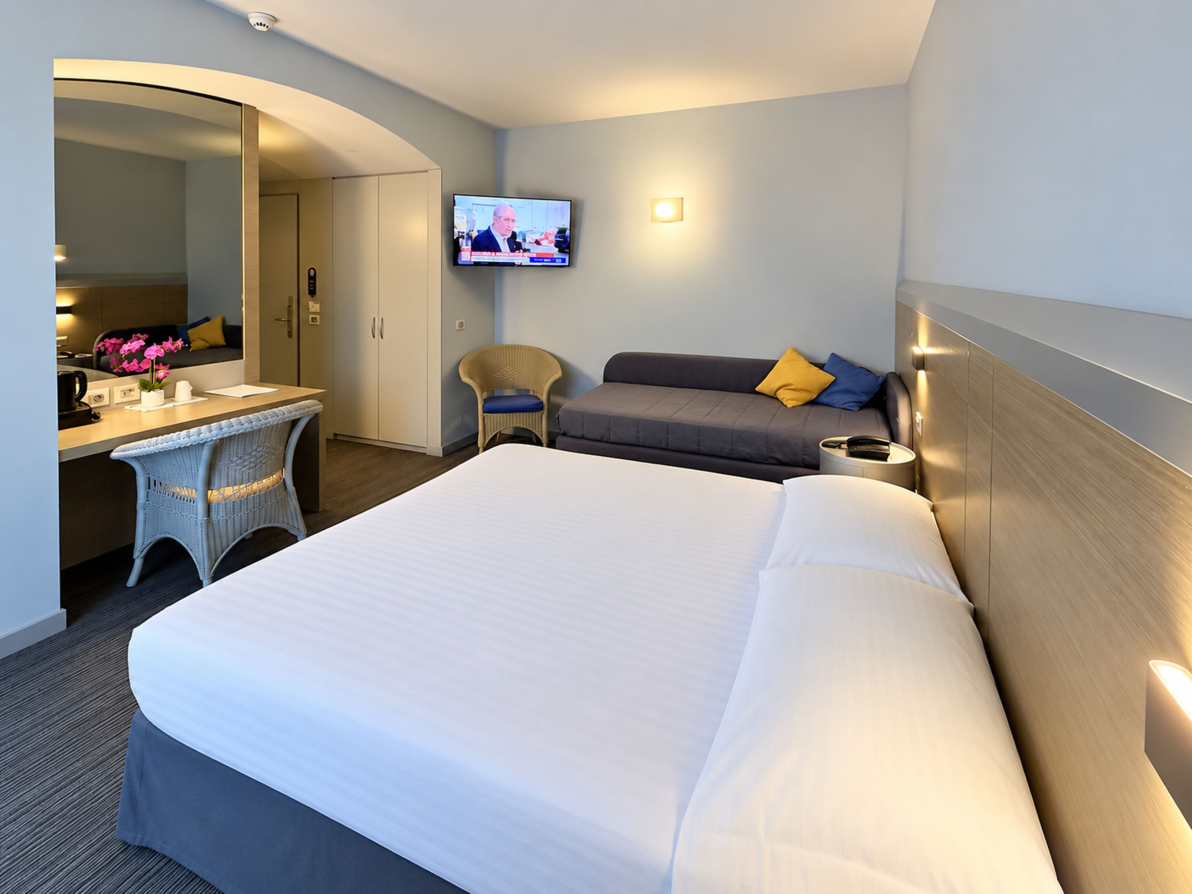 Quarto de hotel com uma cama grande, uma escrivaninha com cadeira, uma televisão na parede e um sofá.