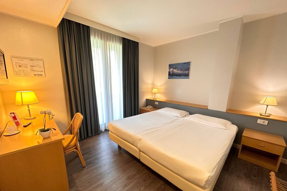A hotel room with a double bed, a desk with a lamp, a chair, a nightstand with a lamp, and a window with curtains.