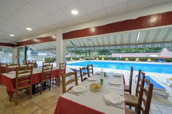 Comedor con mesas dispuestas para el servicio, con vistas a una piscina con sillones y sombrillas.