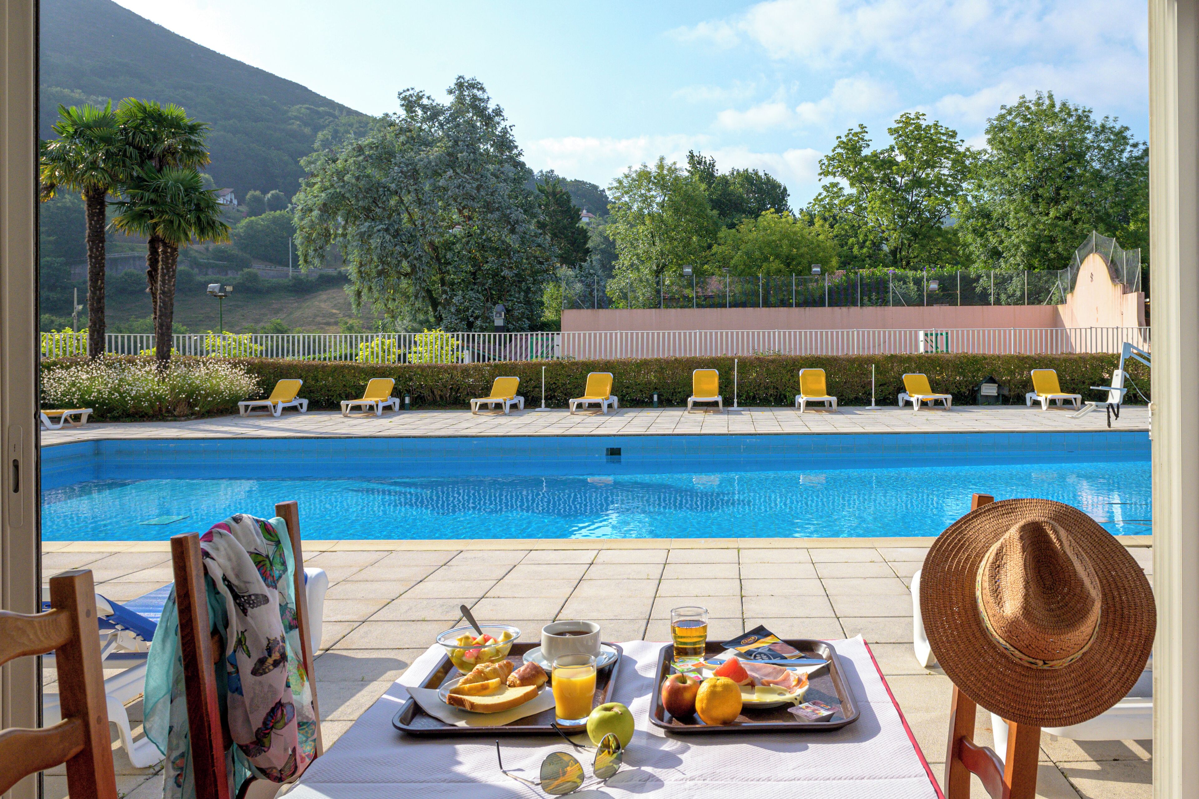A poolside breakfast table with a tray of food, a straw hat, and a view of the pool and surrounding greenery.