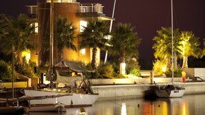 A marina at night with a sailboat docked in the foreground, a building with a lighted upper level, and palm trees along the waterfront. - Agave Apartments (Pisticci)