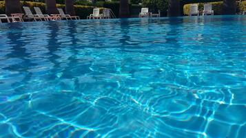 Piscina límpida com água refletindo a luz do sol, rodeada por espreguiçadeiras e palmeiras.