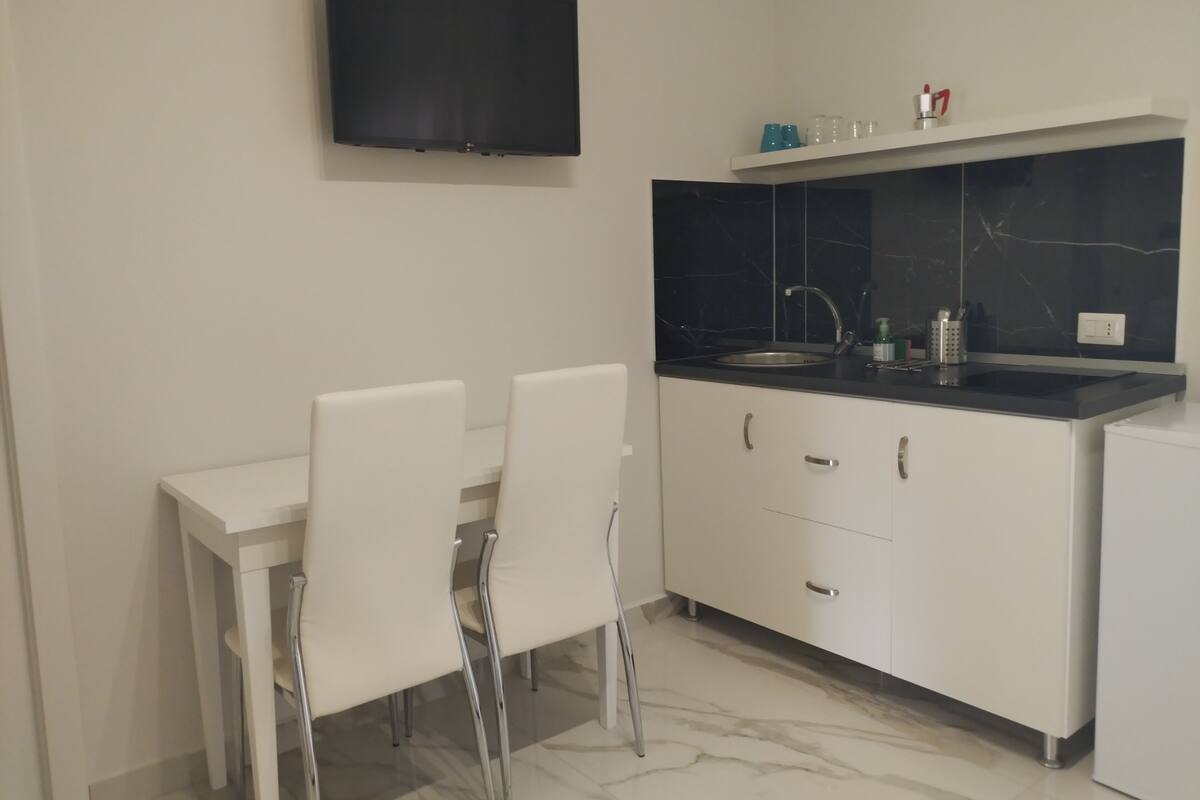 A modern kitchen with a flat-screen TV mounted on the wall, a white dining table with two chairs, a black countertop, and a white refrigerator.