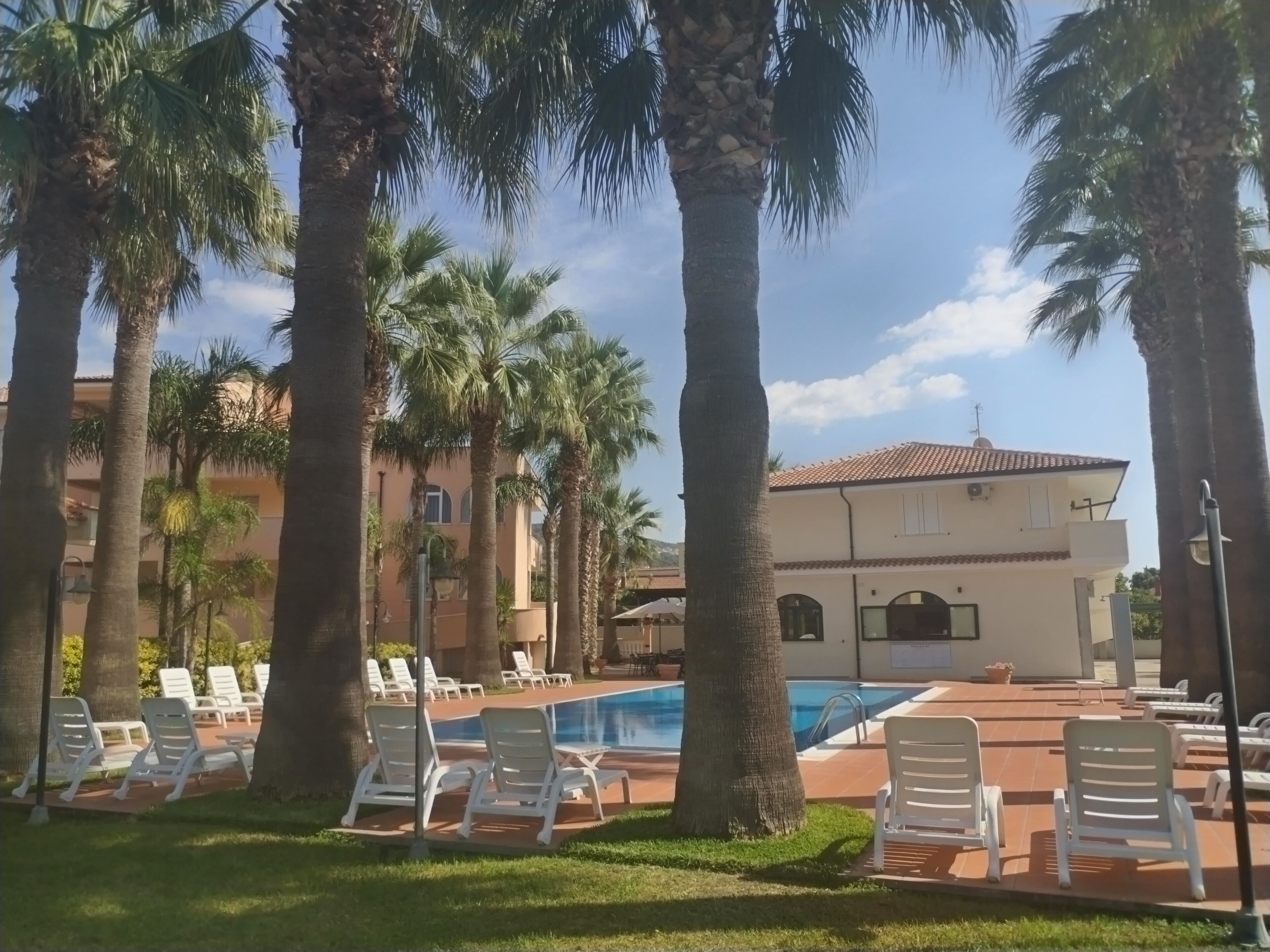 Zona de piscina amb gandules i palmeres, piscina i edifici amb teulada enrajolada.