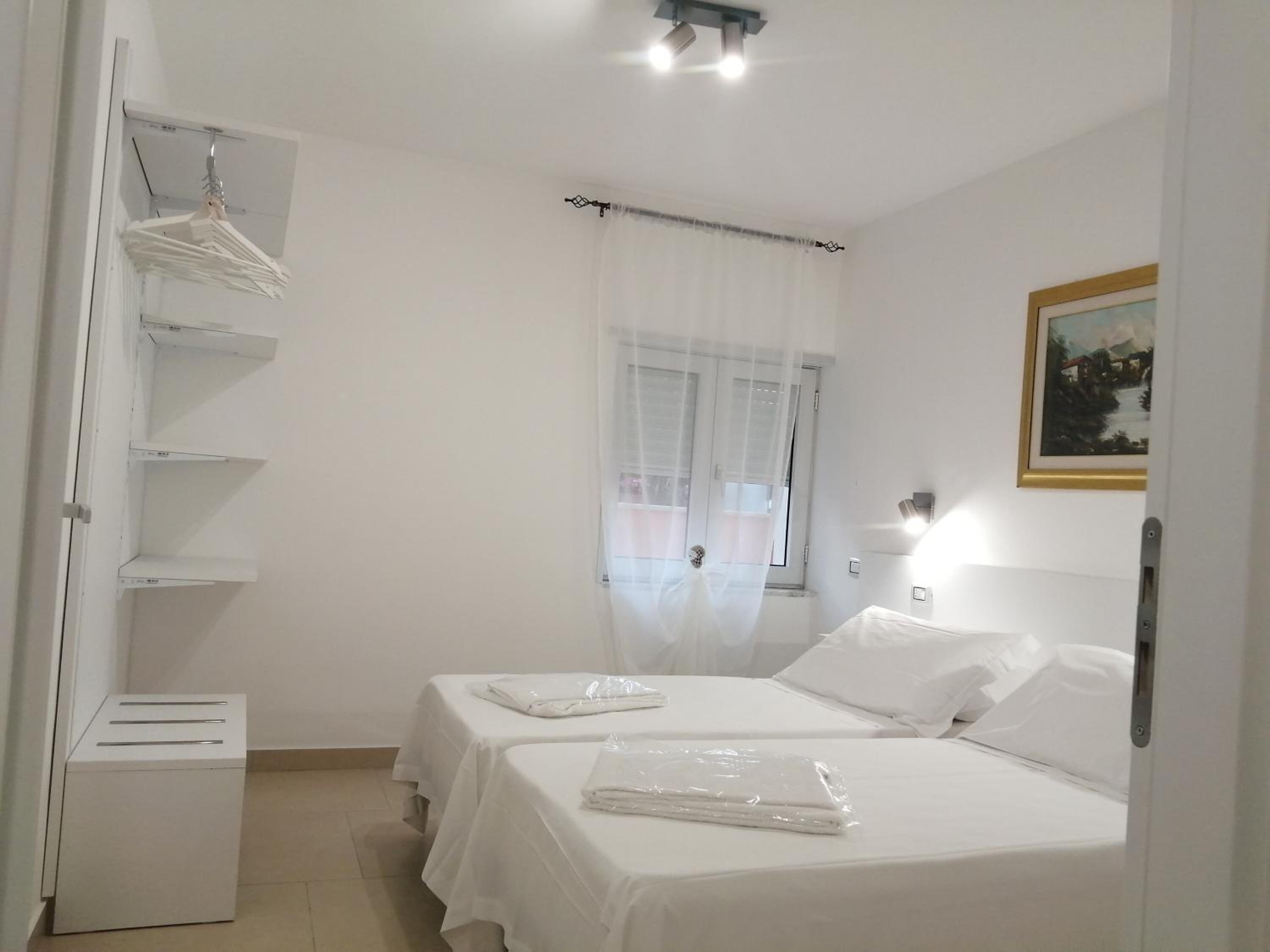 Habitación de hotel con dos camas, un armario con ropa colgada, una mesita de noche, una ventana con cortinas translúcidas y un cuadro en la pared.