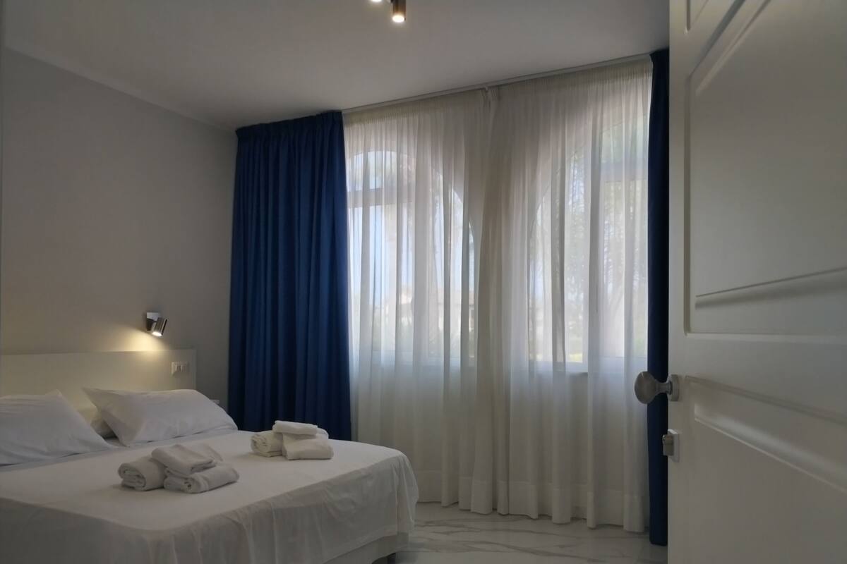 A hotel room with a bed, a nightstand, a wall-mounted lamp, and a window with sheer curtains.