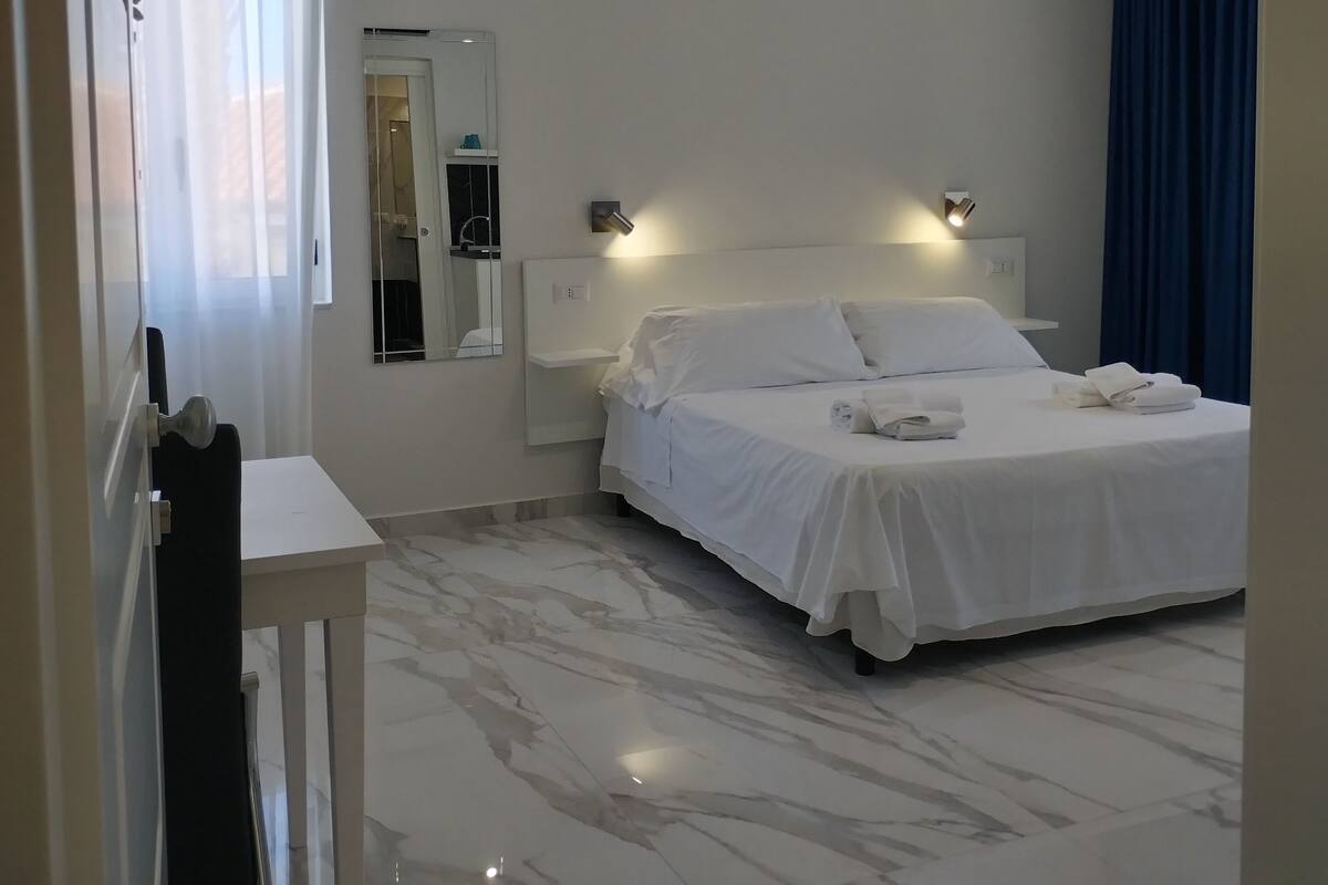 A hotel room with a bed, a small table, a mirror, and a ceiling light fixture.