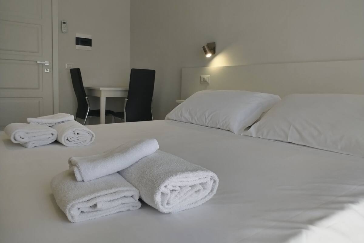 A neatly made bed with white linens, a chair, a small table, and a wall-mounted light fixture.