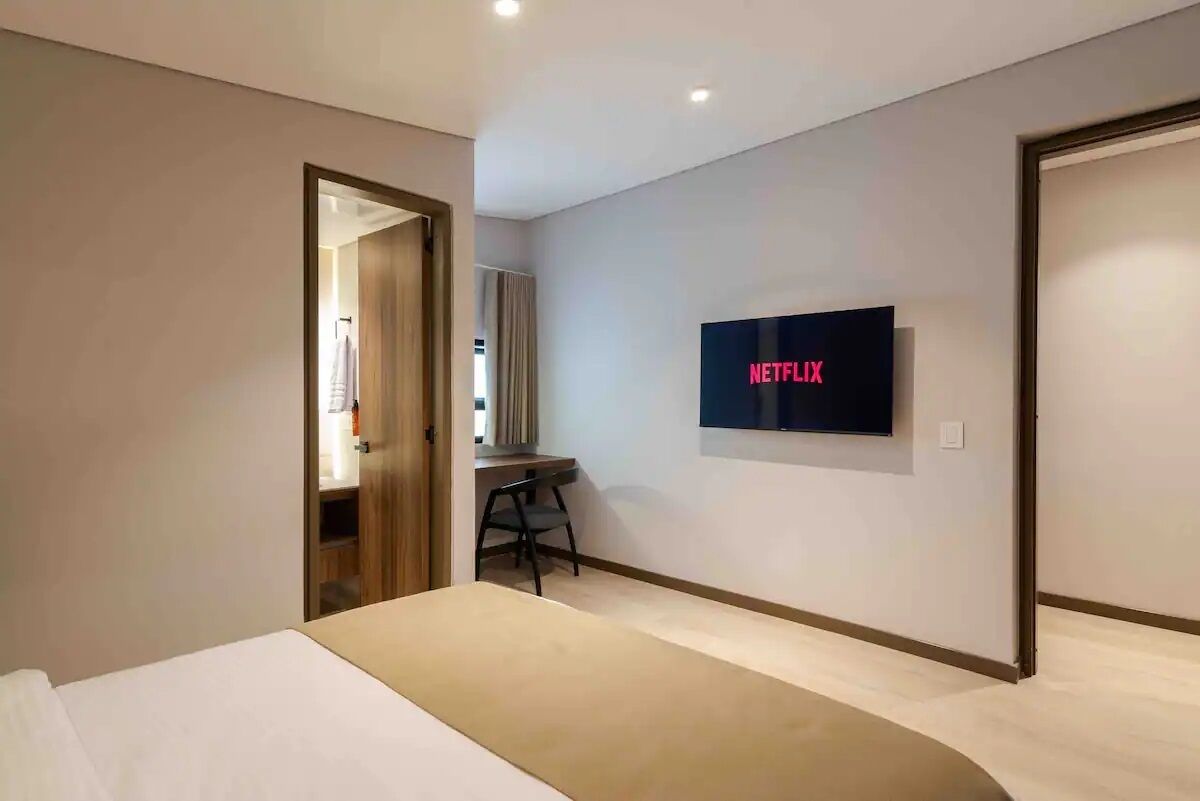 A modern hotel room with a bed, a desk, and a wall-mounted TV displaying the Netflix logo.