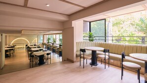 A spacious indoor area with multiple tables and chairs, a seating area with a cushioned bench, and large windows providing natural light and a view of greenery outside. - Hotel Lincoln (Medellín)
