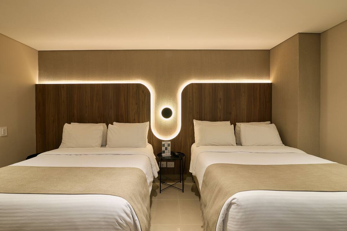 A hotel room with two beds, a nightstand, and a wall-mounted light fixture.