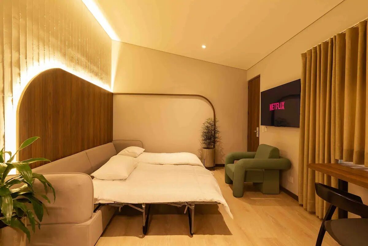 A modern bedroom with a bed, a green chair, a small table, and a TV displaying the Netflix logo.