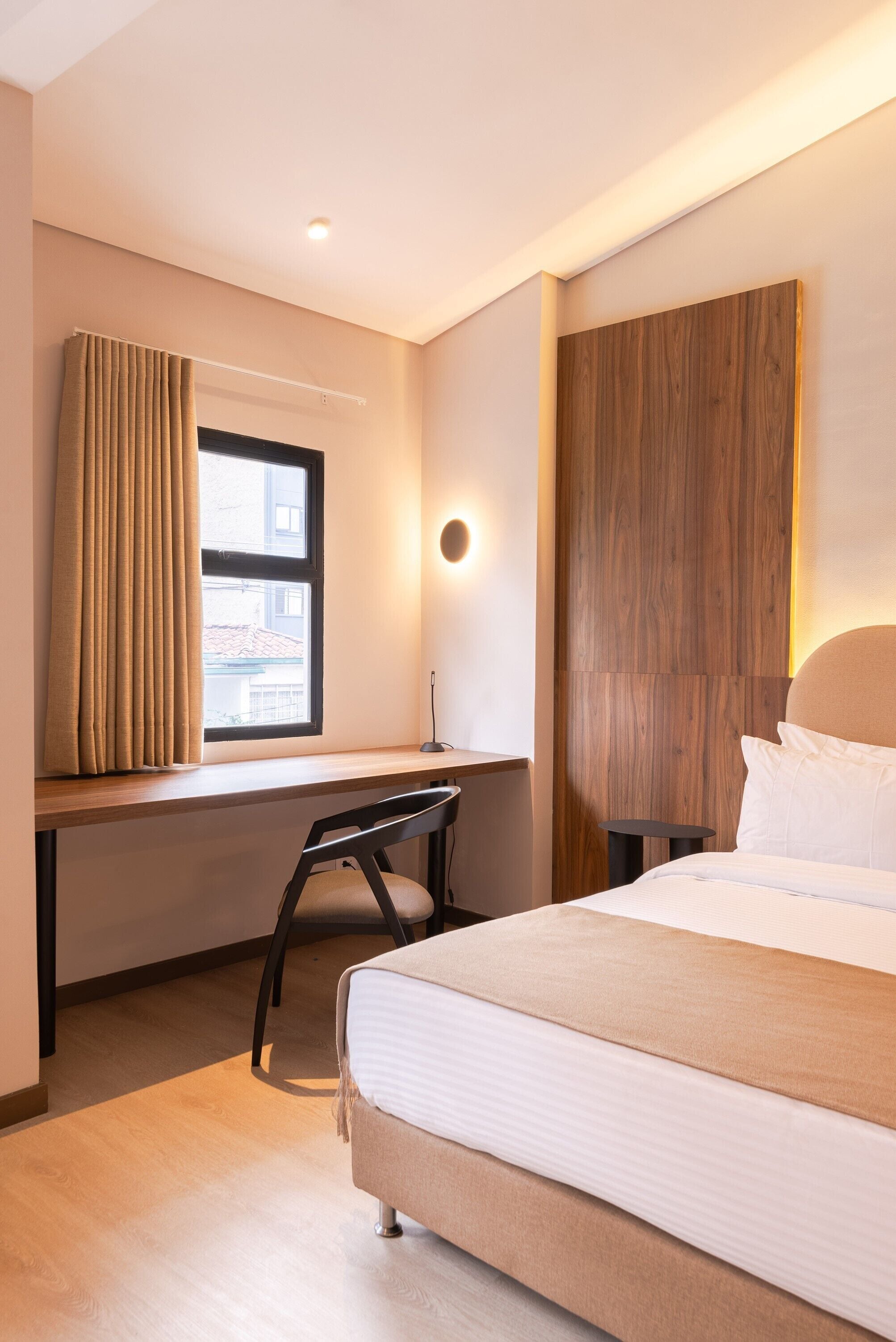 A hotel room with a bed, a desk, a chair, a window with curtains, and a wall-mounted light.