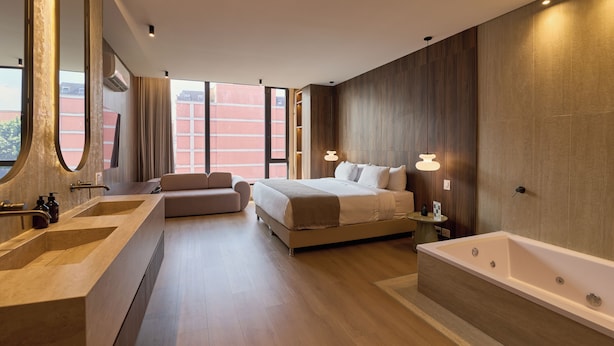 A modern hotel room with a large bed, a separate bathroom with a bathtub, and a large window offering a city view. - Hotel Lincoln (Medellín)