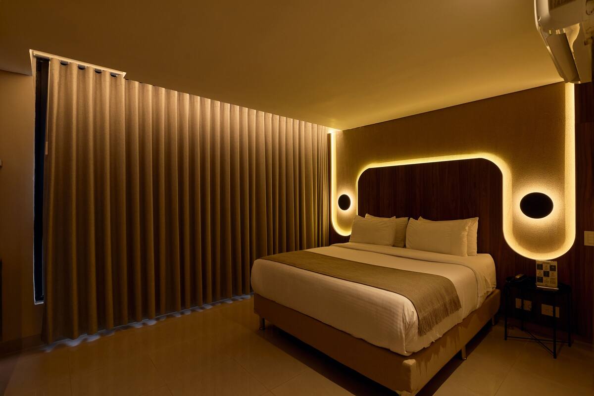 A modern hotel room with a large bed, a wooden headboard, and a wall-mounted light fixture.