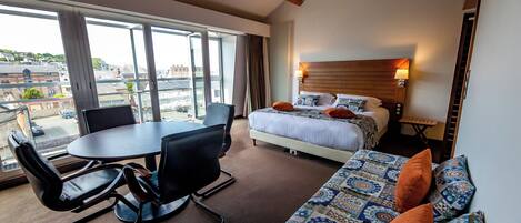 Habitación de hotel con cama, sofá, mesa de comedor con sillas y amplias ventanas con vistas a la ciudad.