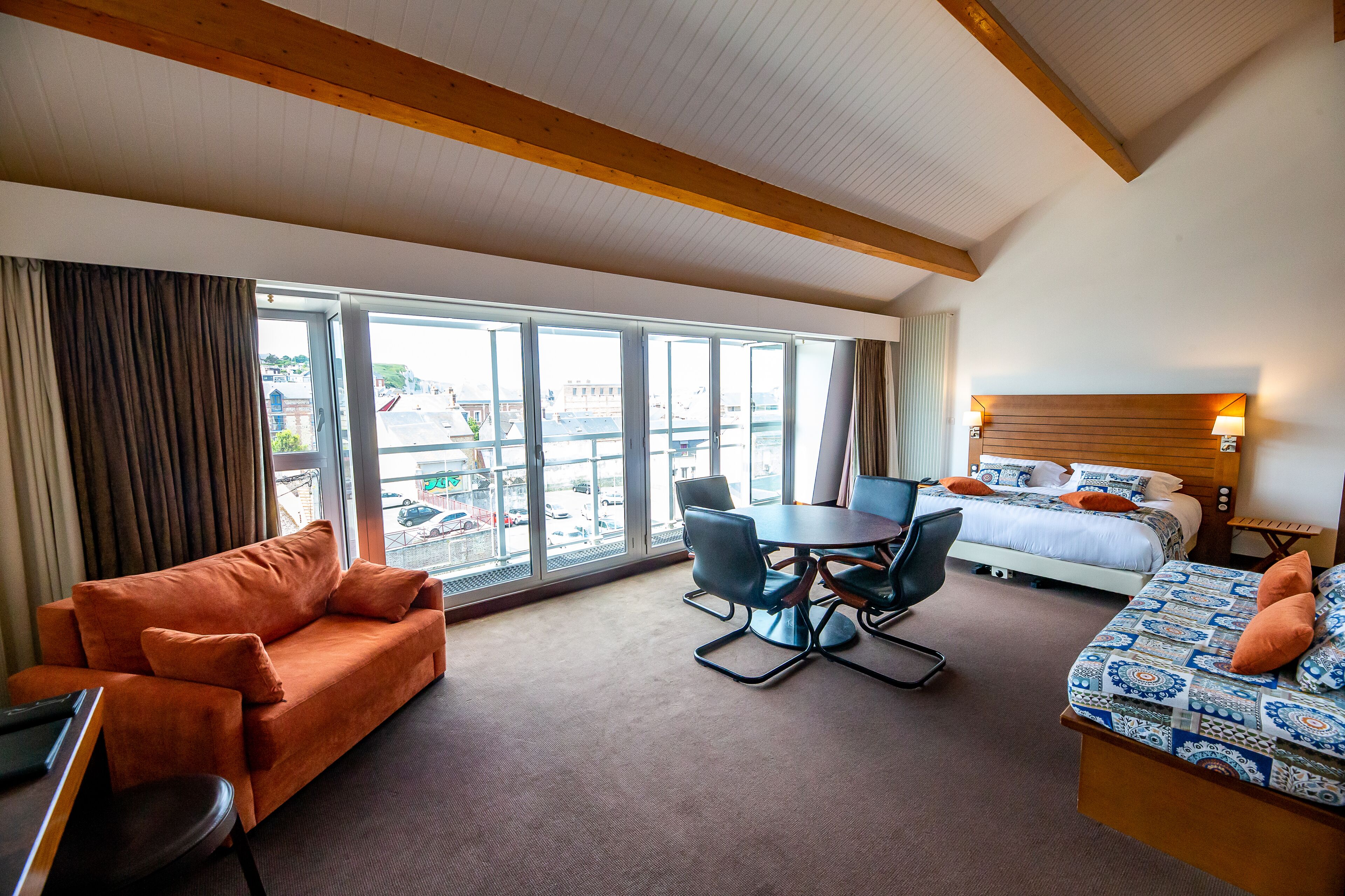 A hotel room with a large window, a bed, a sofa, a dining table, and a desk.