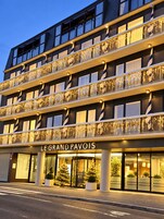 Un hotel de varios pisos con balcones decorados con guirnaldas de luces, una entrada destacada con un letrero que dice 'LE GRAND PAVOIS' y plantas en macetas al frente.