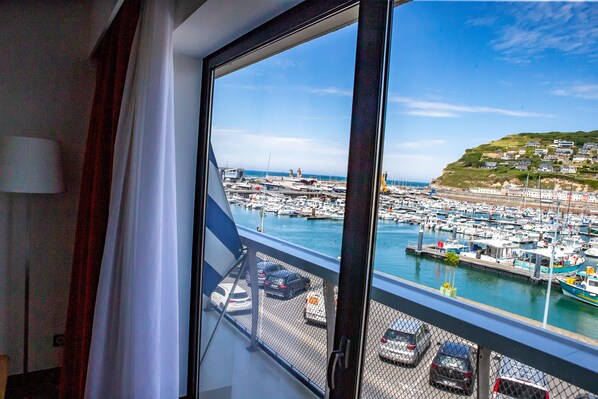 A view from a window overlooking a marina with boats docked, a harbor, and a hillside with buildings. - Le Grand Pavois (Fecamp)