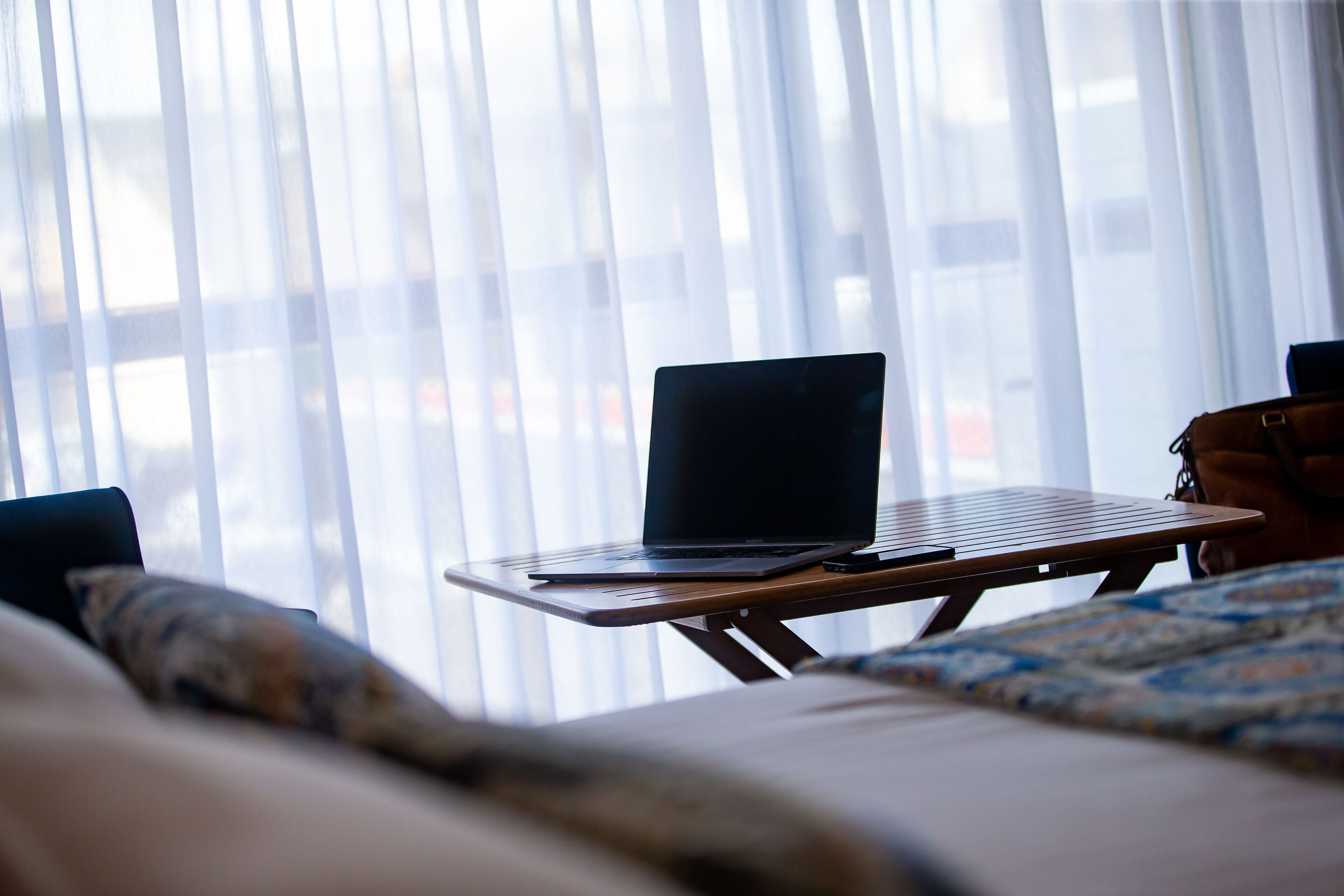 Una laptop sobre una mesa de madera junto a una ventana con cortinas translúcidas.