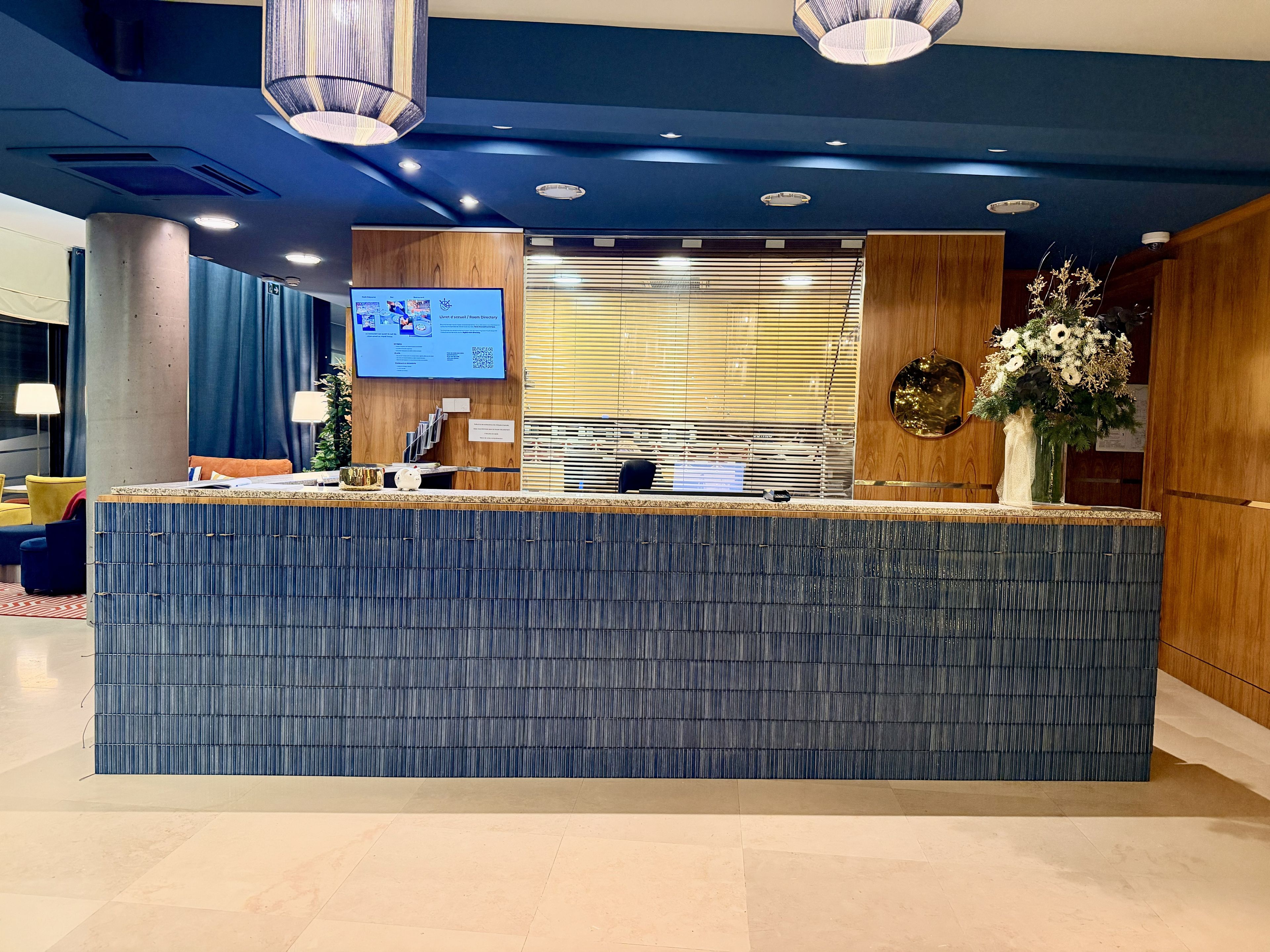 Un mostrador de recepción con pantalla digital, paneles de madera y un jarrón con flores.