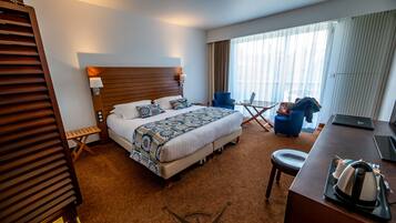 A hotel room with a large bed, a desk with a laptop, a chair, and a view of the outdoors through large windows.