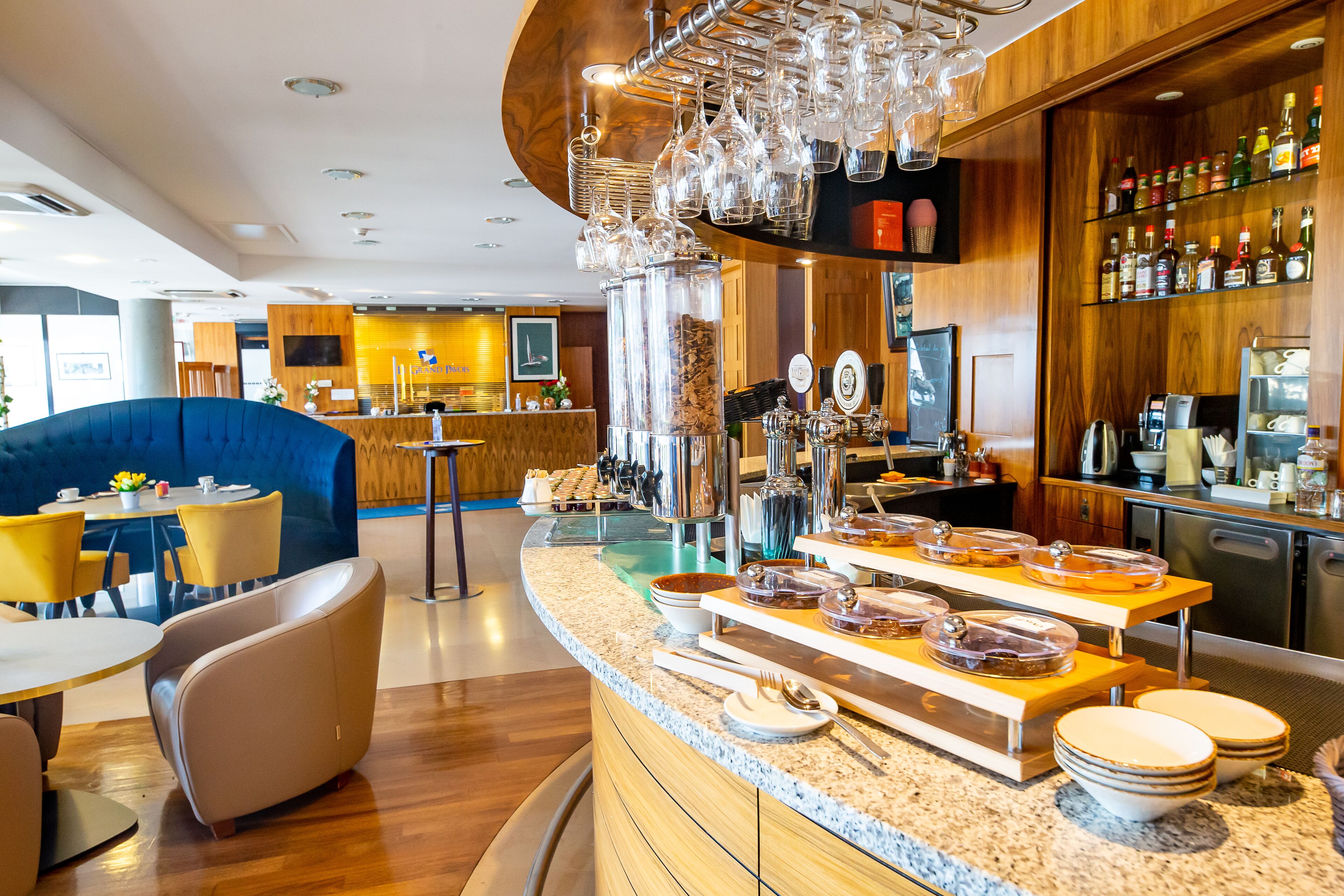 Eine moderne Bar mit einer Theke, Gläsern und einer Auswahl an Flaschen. Es gibt eine Sitzecke mit einem blauen Sofa und gelben Stühlen.