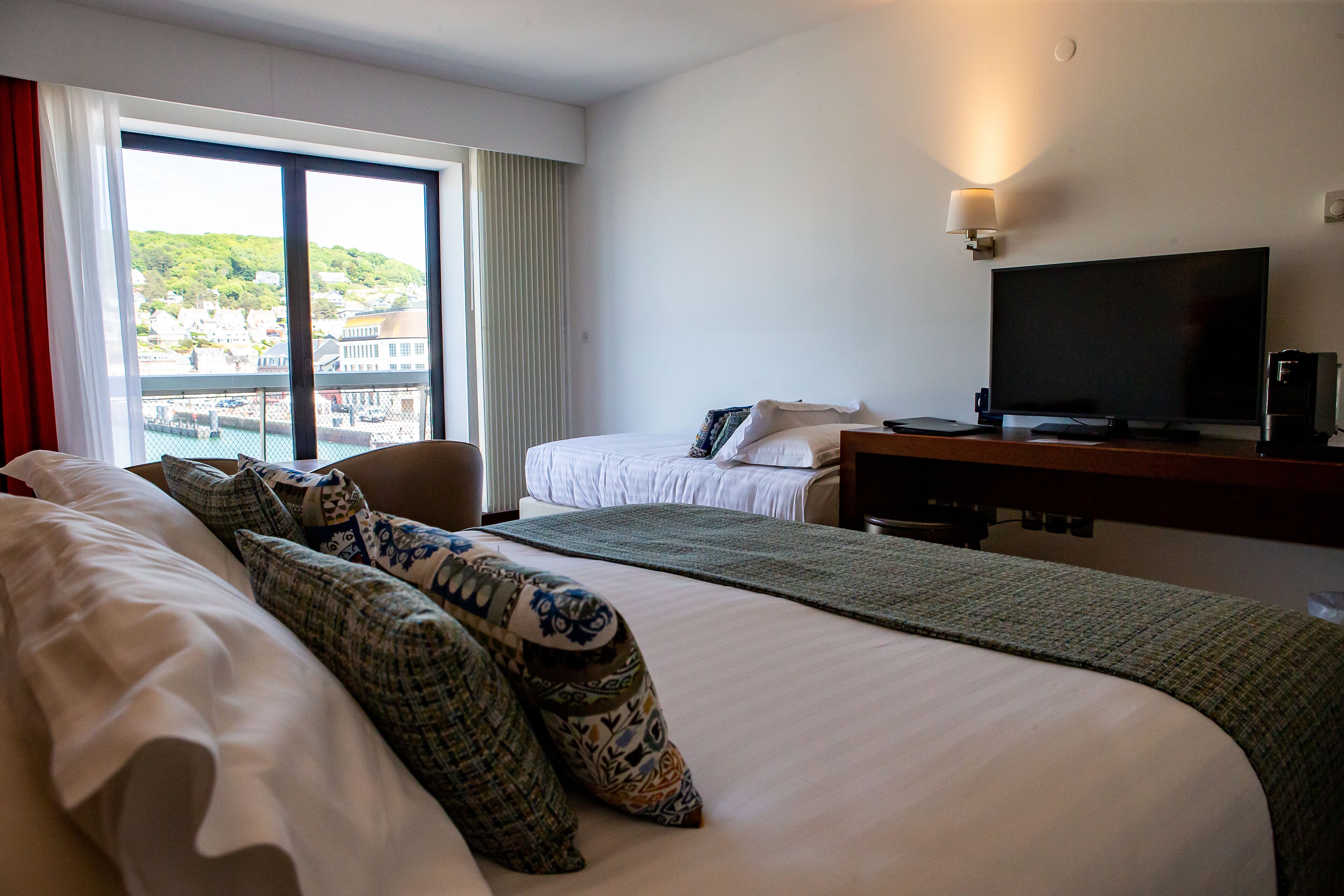 A hotel room with a large bed, a smaller bed, a desk with a TV, and a view of a town through large windows.