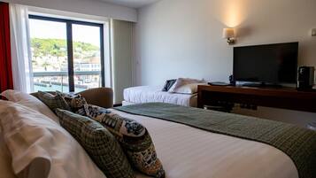 A hotel room with a large bed, a smaller bed, a desk with a TV, and a view of a town through large windows.