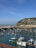A marina with boats docked, a lighthouse in the background, and a hillside with buildings.
