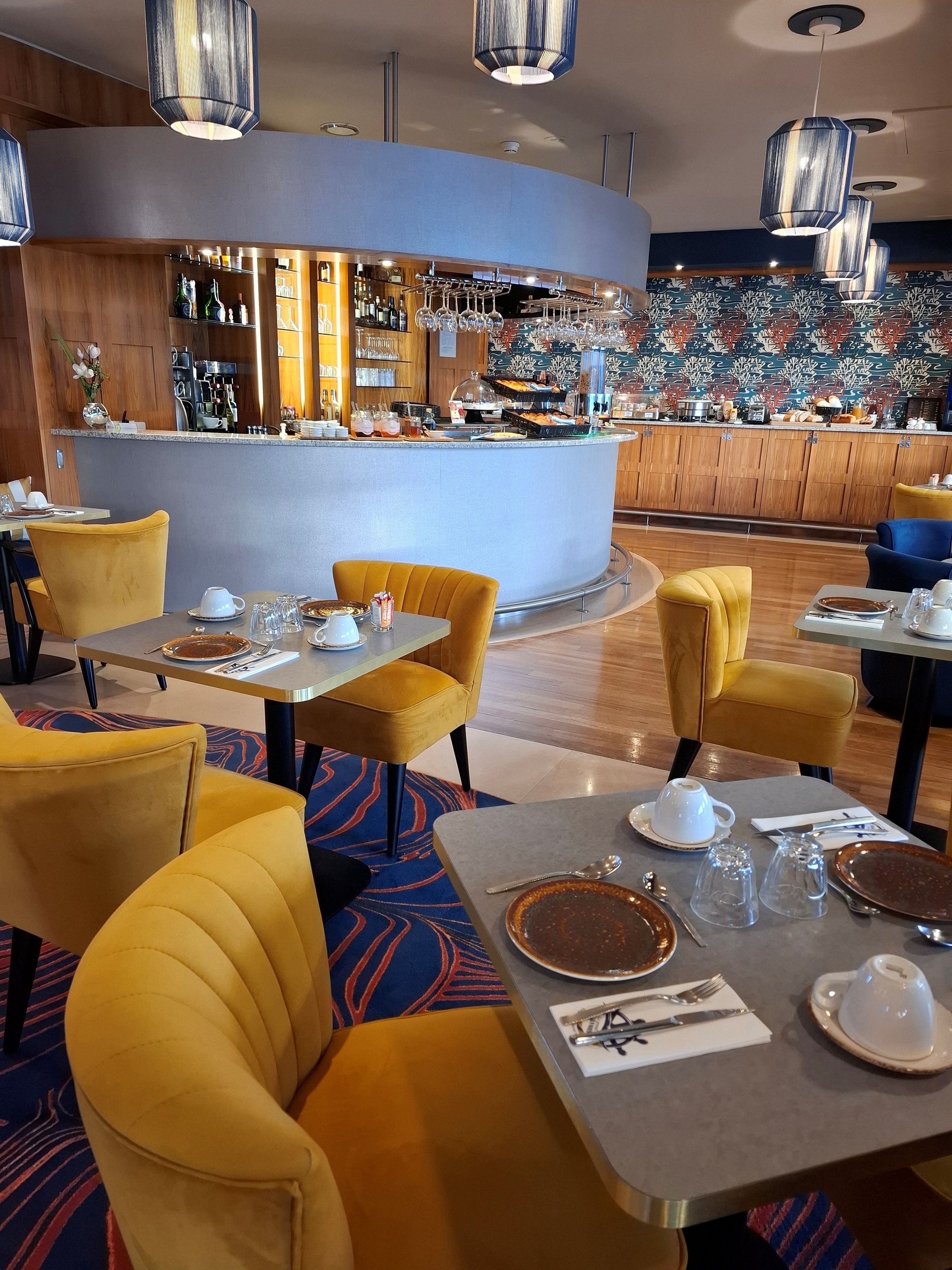 Een restaurant met gele bekleden stoelen, een bar met diverse drankjes en een decoratieve muur op de achtergrond.