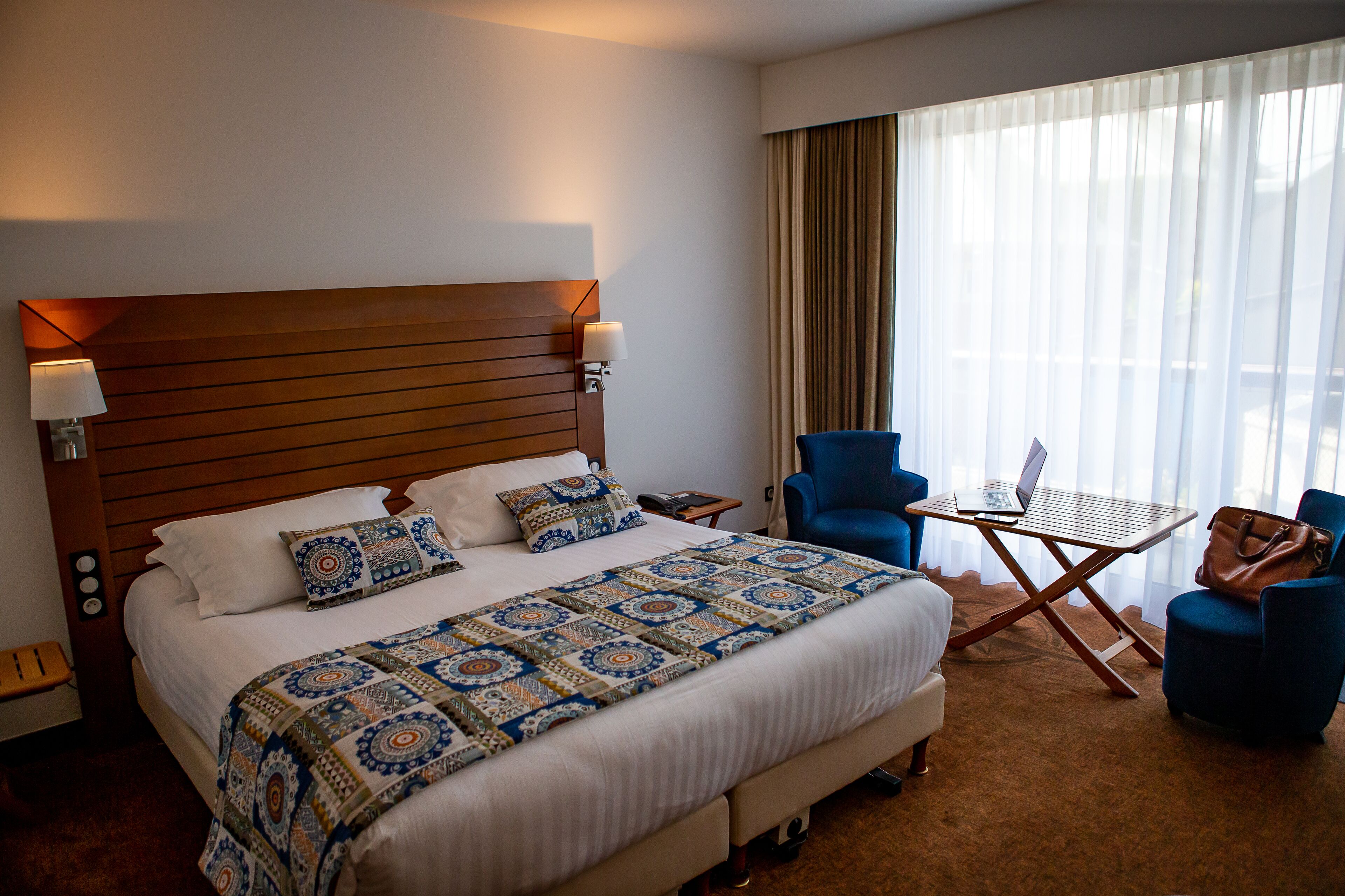 Ein Hotelzimmer mit einem großen Bett, einem Schreibtisch mit Laptop, zwei Stühlen und einem Fenster mit Vorhängen.