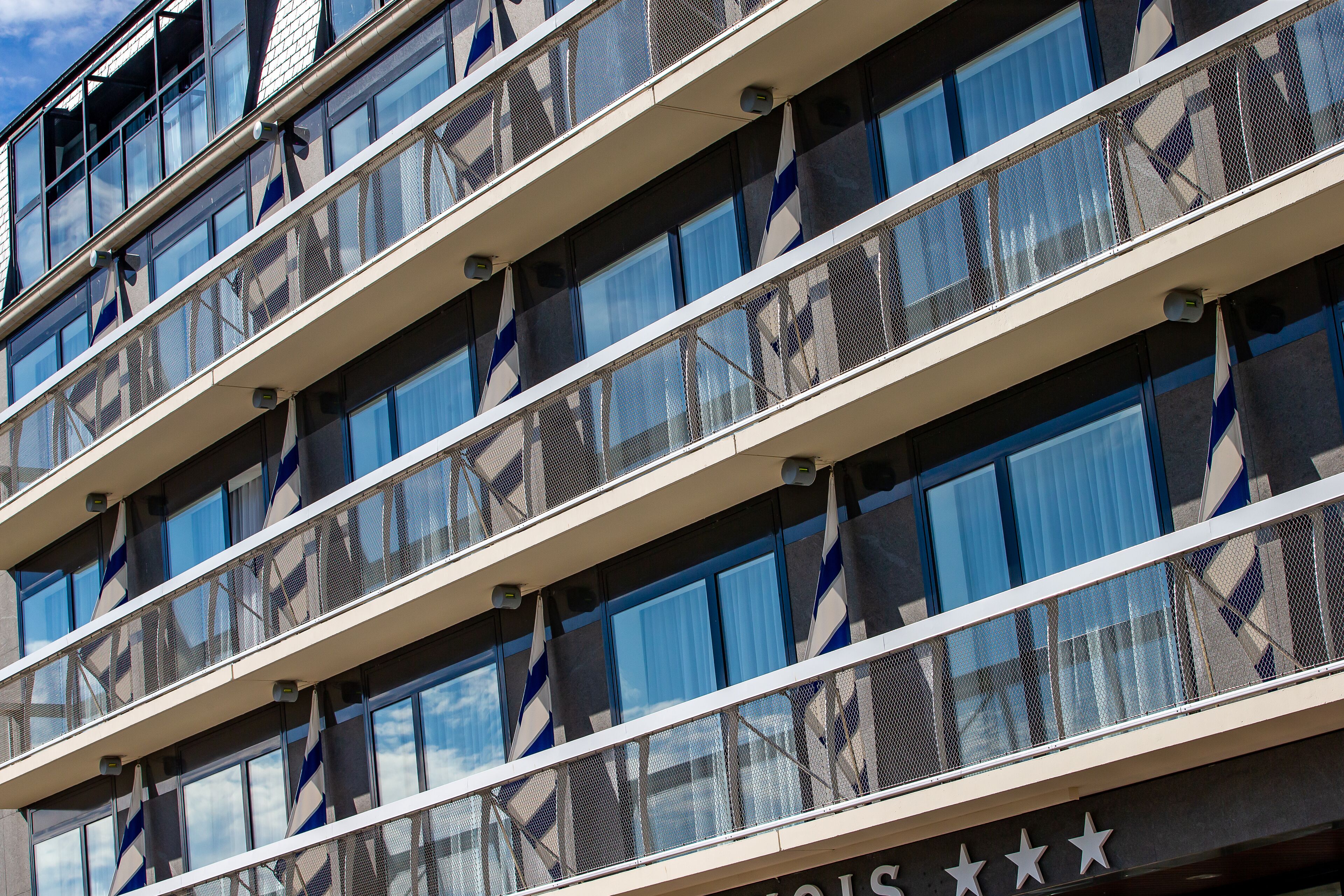 A multi-story building with a modern architectural design, featuring a series of balconies with mesh railings and a clear view of the sky through the windows.