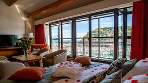 A hotel room with a large window offering a view of a marina and hills. There is a bed with rose petals, a TV, and a table with a vase of flowers. - Le Grand Pavois (Fecamp)