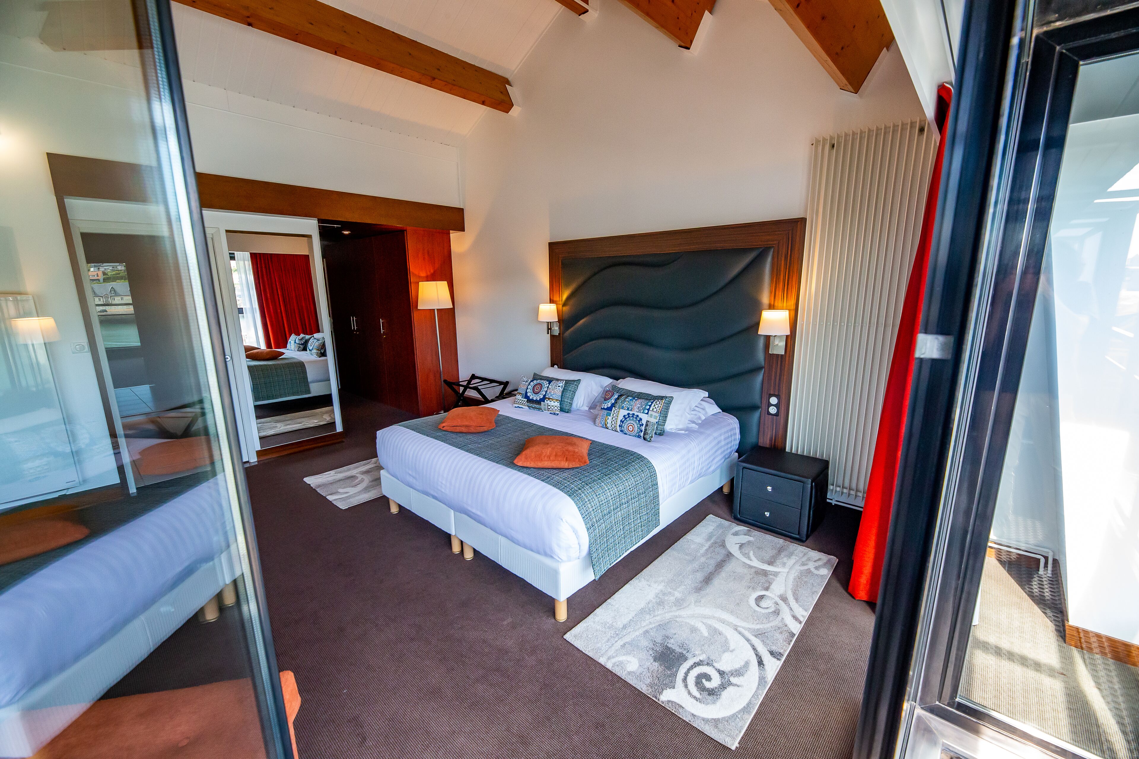 A modern hotel room with a large bed, a wooden headboard, and a glass door leading to a balcony.