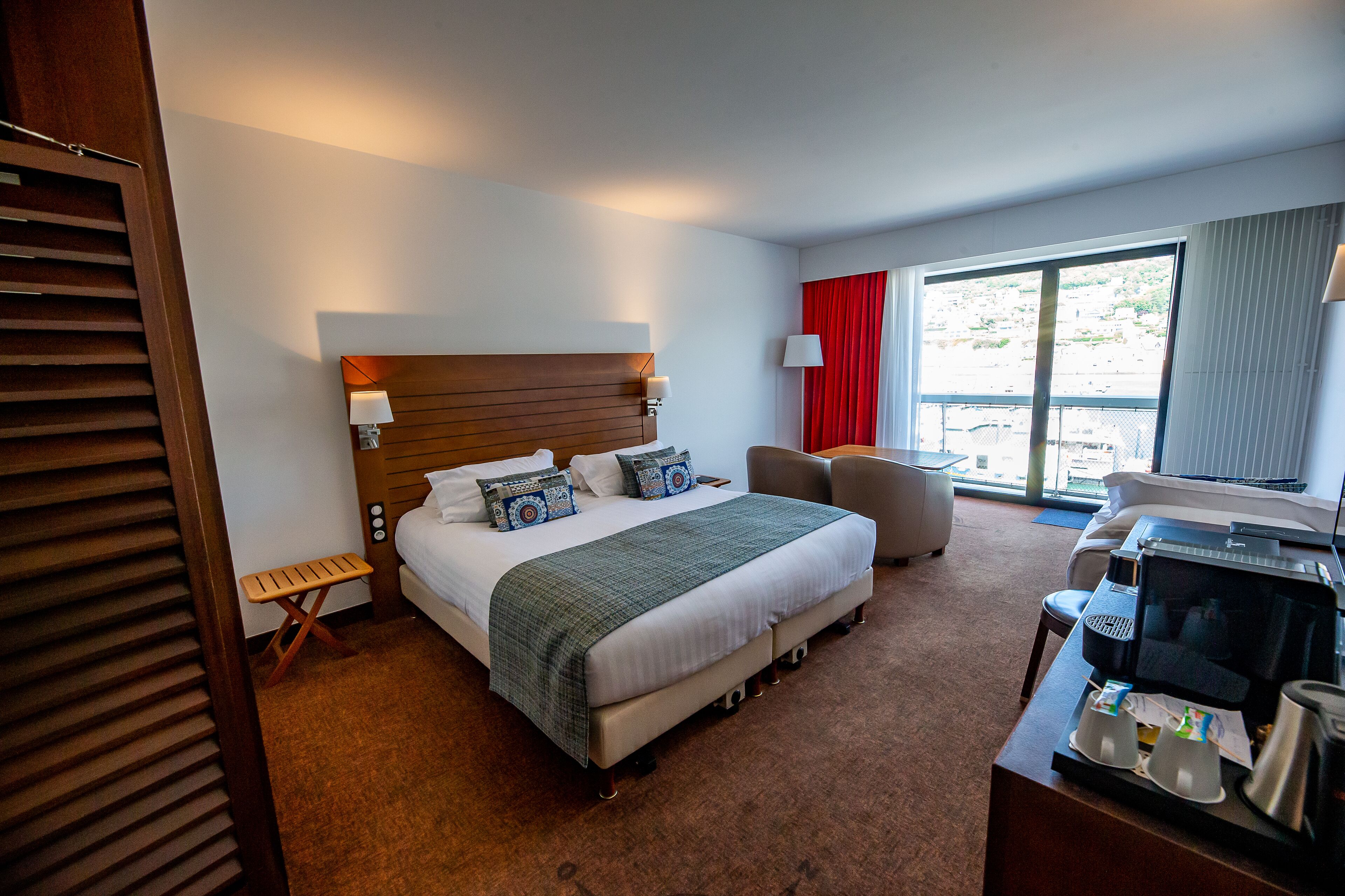A hotel room with a large bed, a desk with a computer, a chair, and a window with curtains.