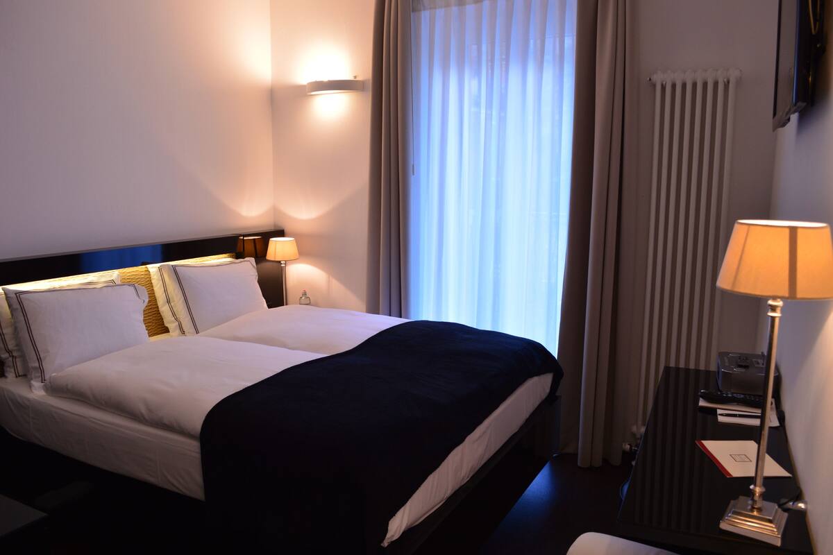 A hotel room with a bed, two bedside lamps, a nightstand, and a window with curtains.