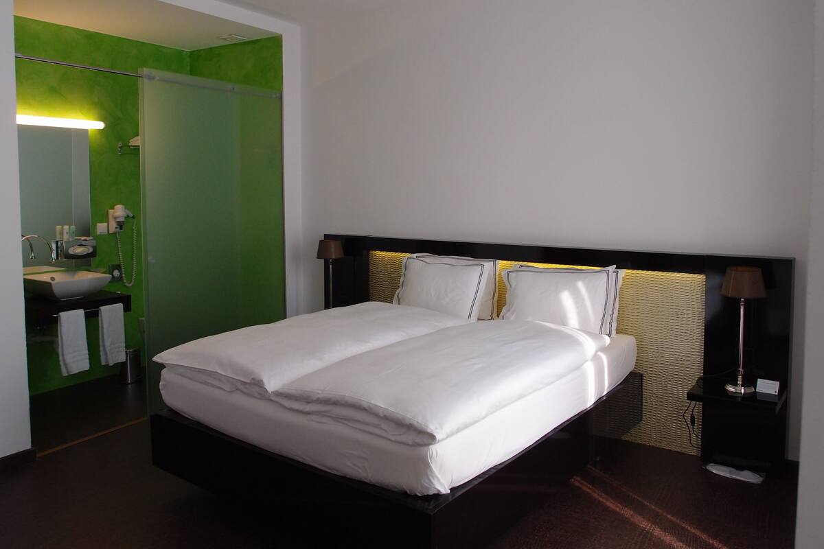 A hotel room with a bed, a bedside table, a lamp, a mirror, and a bathroom with a sink and towel rack.
