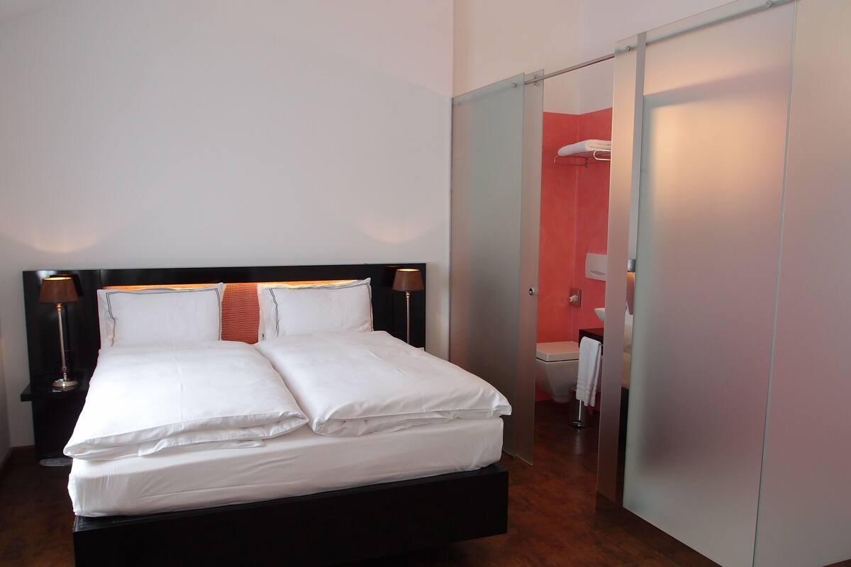A modern hotel room with a large bed, a nightstand with a lamp, a bathroom with a toilet and sink, and a sliding door closet.
