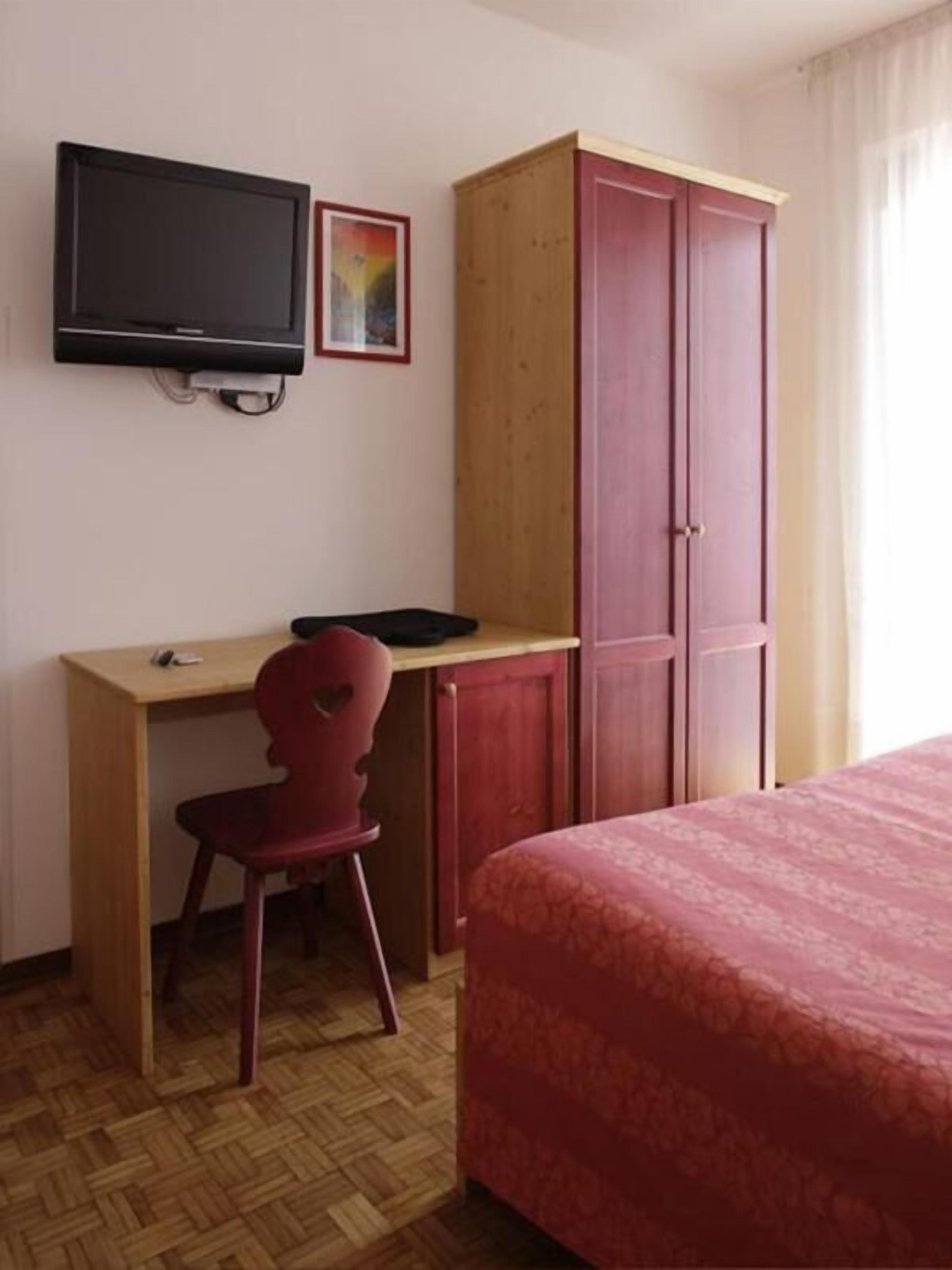 Una habitación con cama, escritorio y silla, televisor montado en la pared y armario.