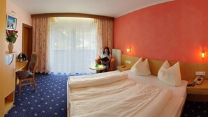 A hotel room with a bed, a desk, a chair, a small table, and a window with curtains. - Hubertus (Fuegenberg)
