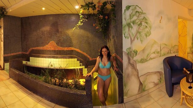 A spa area with a hot tub, a mural of a mountainous landscape, and a seating area with two chairs and a small table. - Hubertus (Fuegenberg)