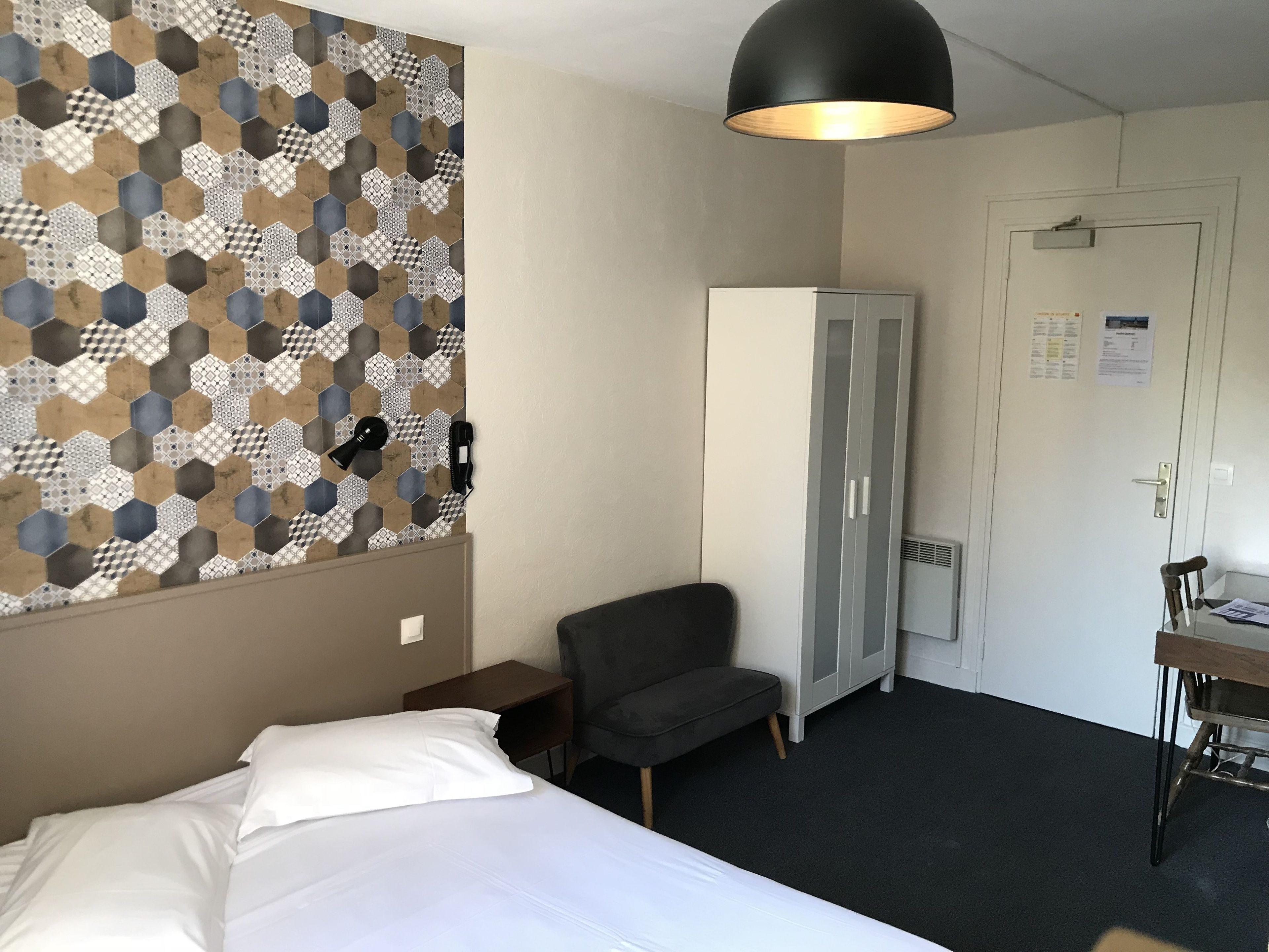 Une chambre d’hôtel avec un lit, un fauteuil gris, une armoire et un bureau avec une chaise.