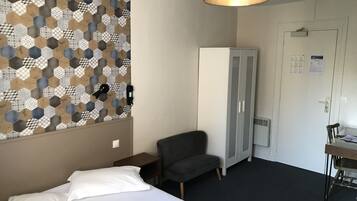 Une chambre d’hôtel avec un lit, un fauteuil gris, une armoire et un bureau avec une chaise.
