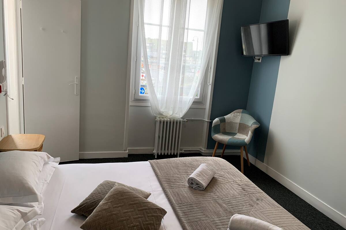 A hotel room with a bed, pillows, a chair, a TV, and a window with curtains.