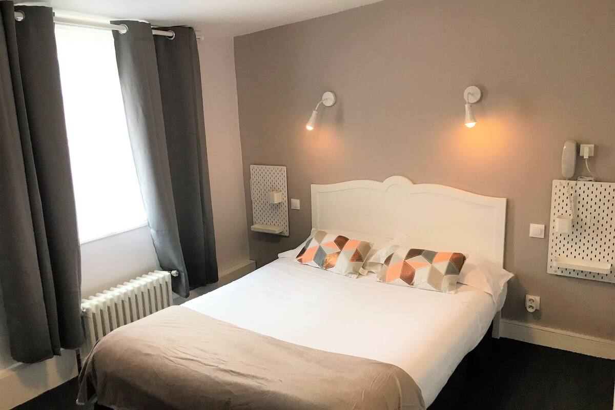A hotel room with a bed, curtains, a radiator, wall-mounted light fixtures, and a wall socket.